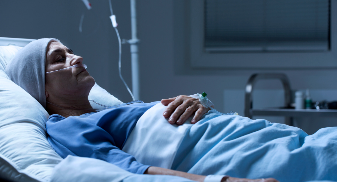 An older woman lies in a hospital bed with an oxygen tube in her nose, wearing a headscarf and hospital gown, covered by a blanket, in a dimly lit Miami Dade medical room.