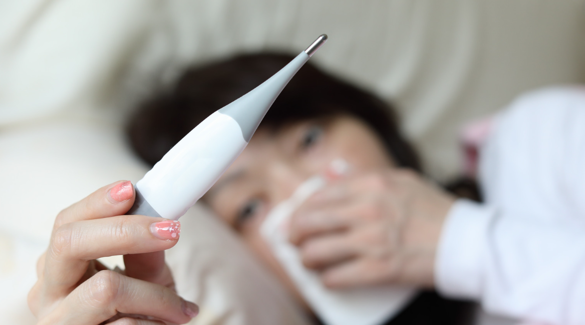 A person lies in bed holding a digital thermometer in the foreground, with their other hand holding a tissue to their nose, suggesting they are unwell—perhaps considering pest control services in Miami to help ensure a healthier home environment.