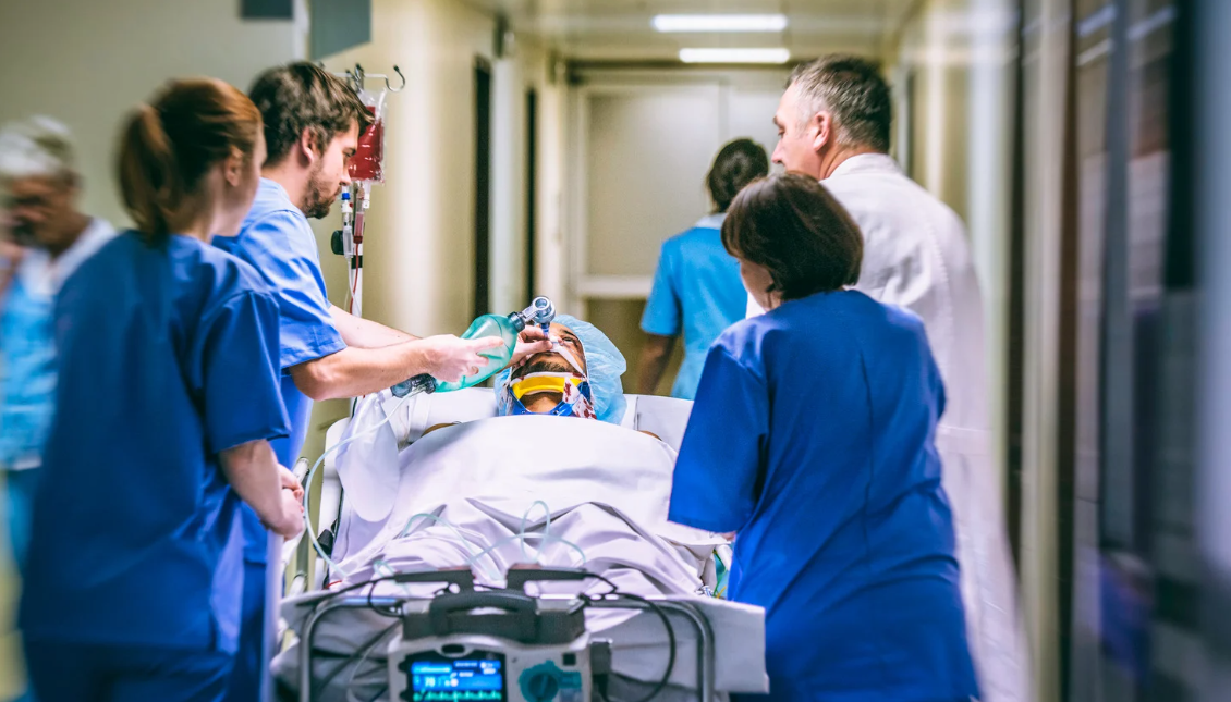 A group of medical staff in scrubs quickly wheel a patient on a hospital stretcher down a hallway in Miami; one person holds an oxygen mask to the patient’s face.