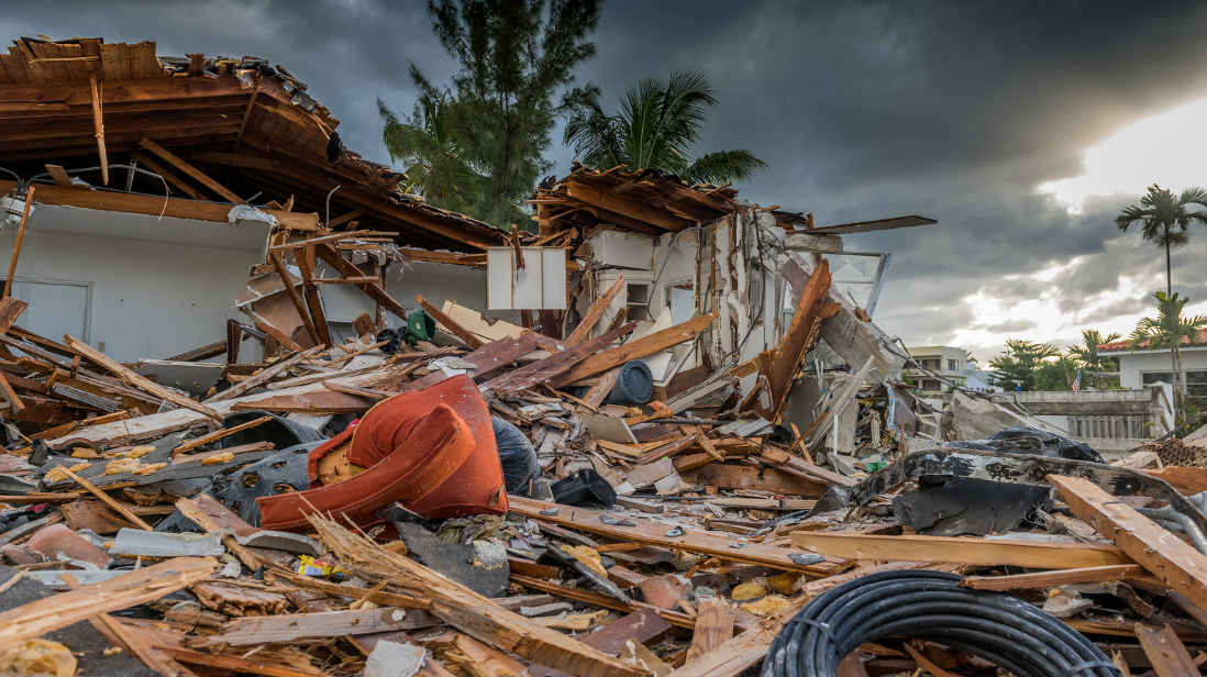 A destroyed house with debris, broken wood, and household items scattered everywhere; a red chair is visible among the wreckage under a dark, cloudy sky with palm trees in the background, typical of storm-ravaged South Florida.