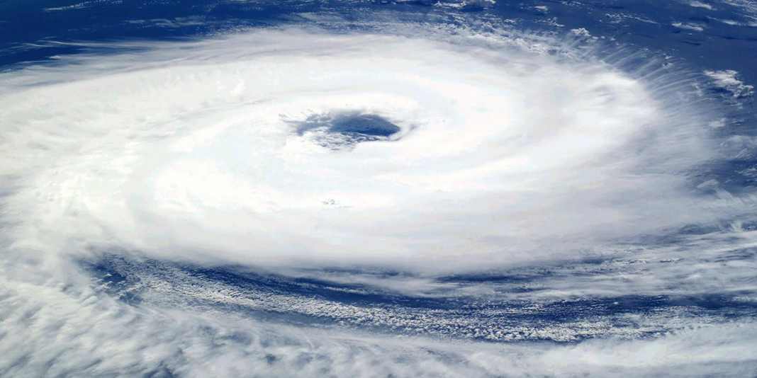 A massive hurricane with a well-defined eye swirls over the ocean near South Florida, seen from above. Thick white clouds spiral outward, contrasting with the deep blue water below.