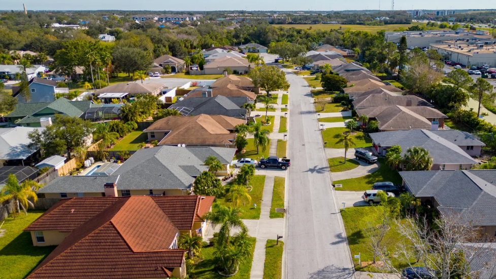 Aerial view of a suburban Miami-Dade neighborhood with single-family houses, green lawns, trees, and cars parked in driveways along a straight residential street under a clear South Florida sky.
