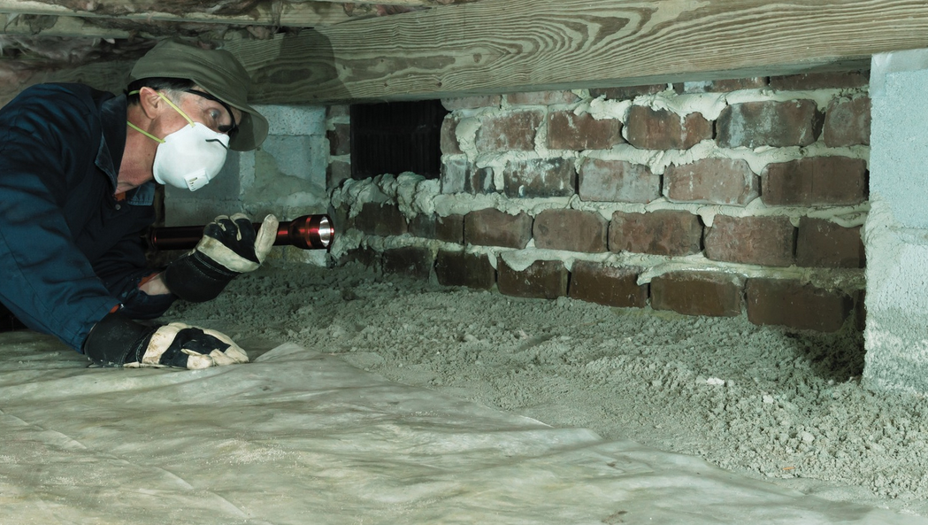 A person wearing a mask, gloves, and a hat inspects a Florida crawl space under a house with a flashlight, examining the dirt floor and brick wall for signs of rats.